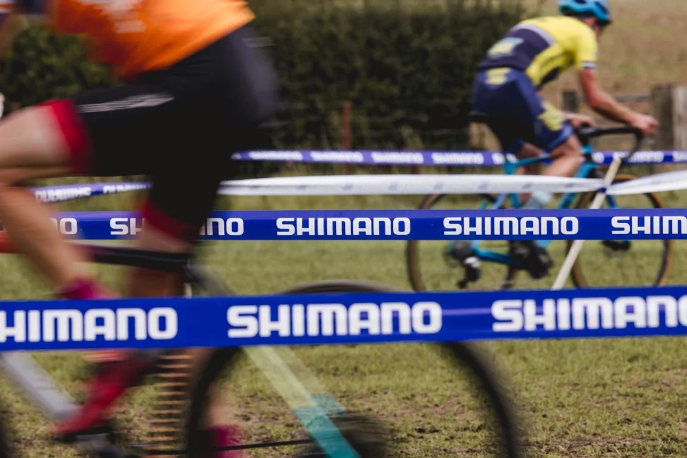 Two cyclists riding fast through a course marked with blue Shimano tape on a grassy field.
