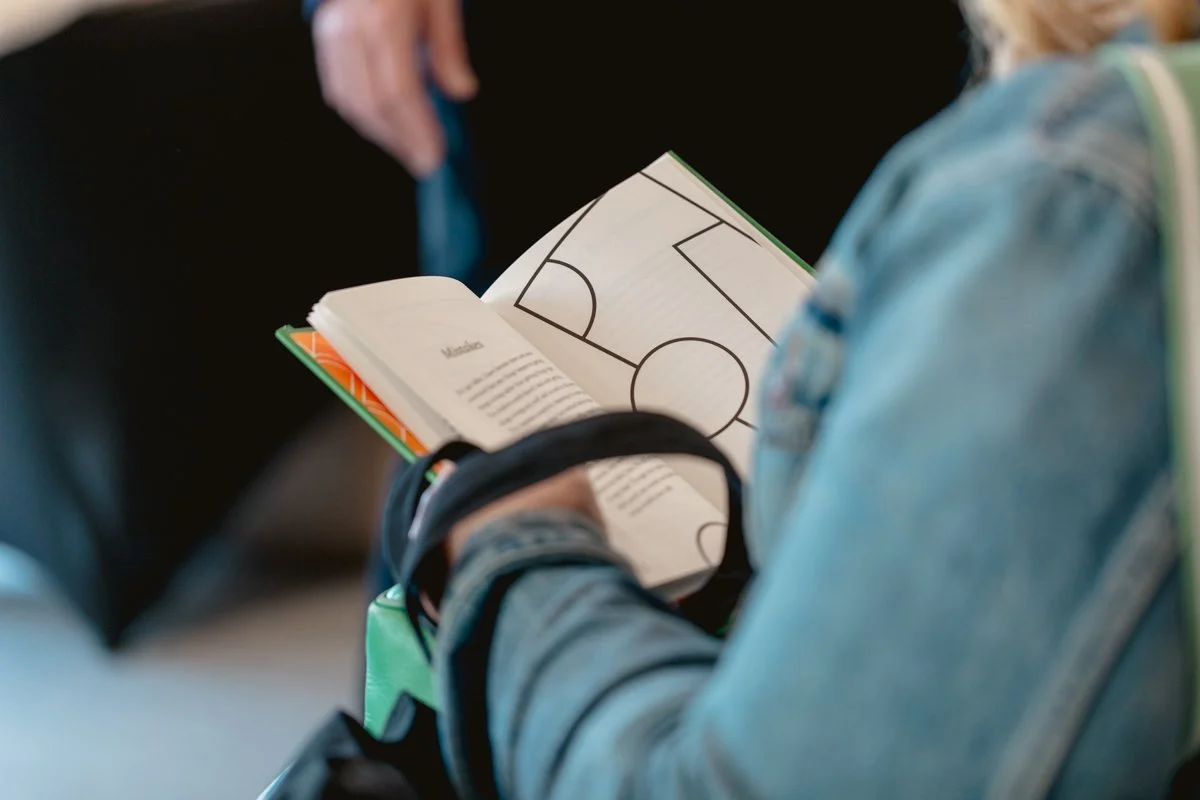 Person reading a book with a soccer field diagram inside, wearing a denim jacket, with a backpack nearby.