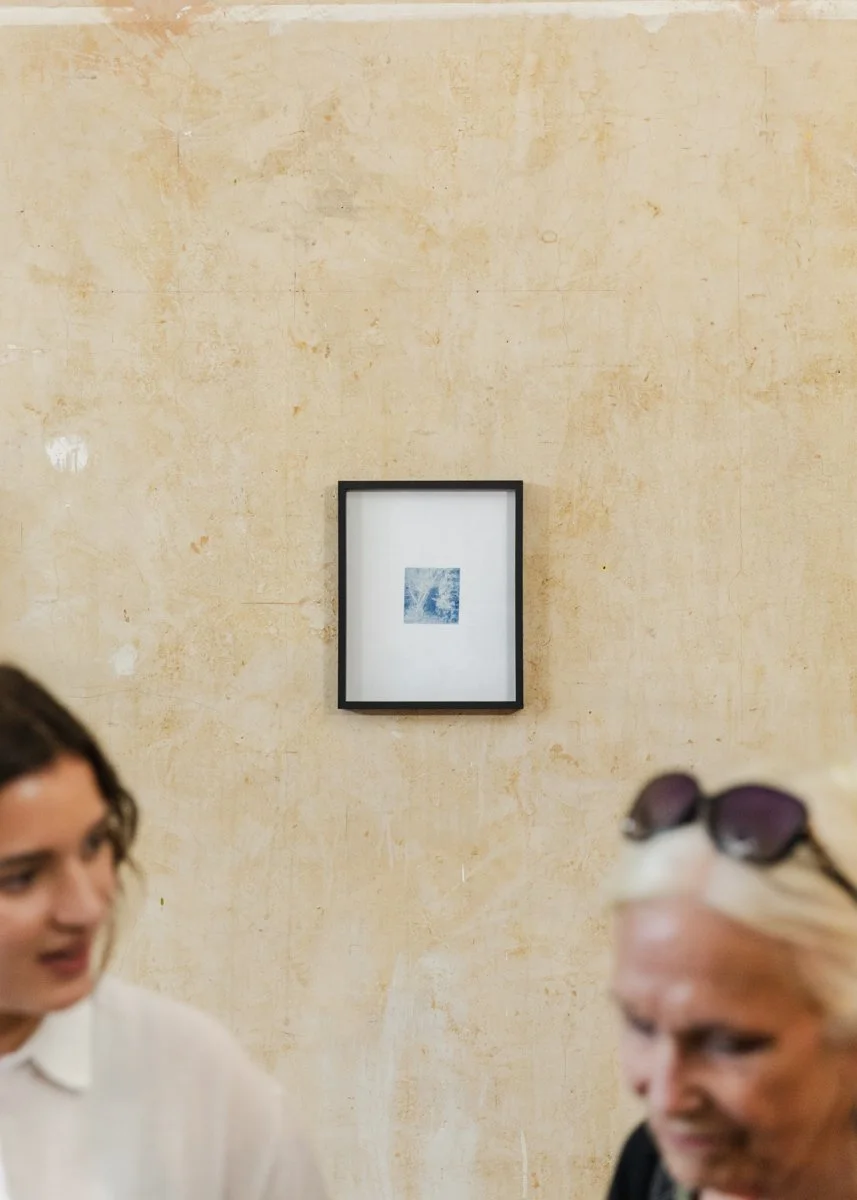Two women having a conversation indoors, with a framed artwork on a beige wall in the background.