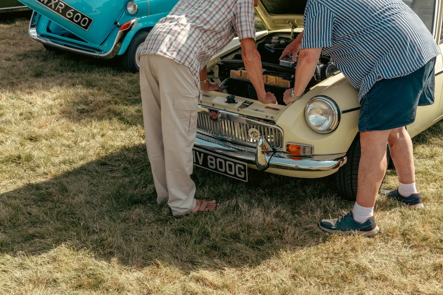 Two men work on a beige vintage MG car with its hood open, parked on grass during daytime, with a blue classic car in the background.