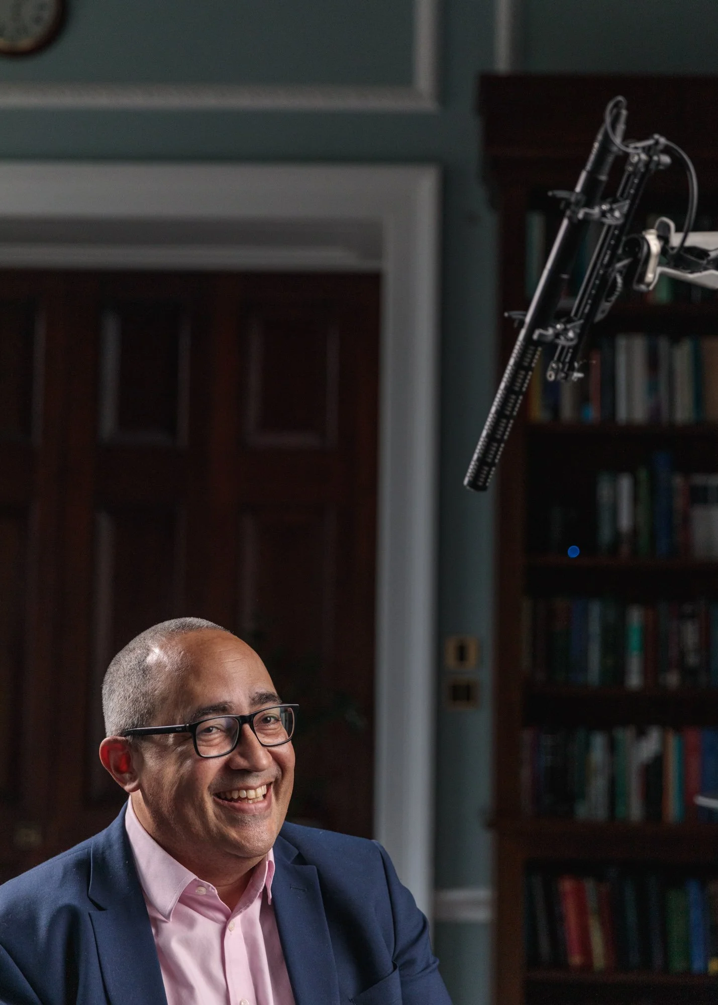 A man with glasses and a pink shirt smiling, sitting at a desk in a room with bookshelves, a wooden door, and a microphone suspended in mid-air.