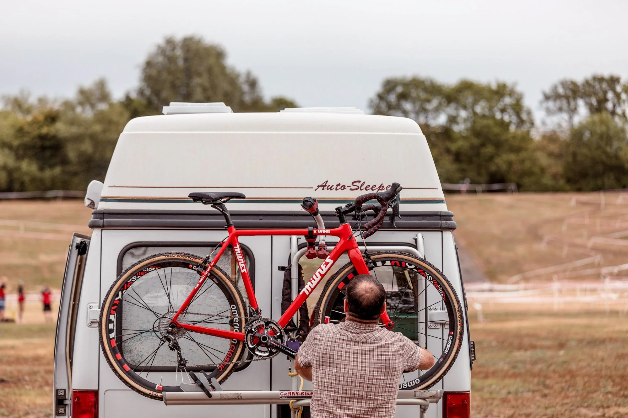 A man standing behind a white camper van, working on a red bicycle mounted on the back of the van. The bicycle is secured on a rack, with a background of open field, trees, and a cloudy sky.