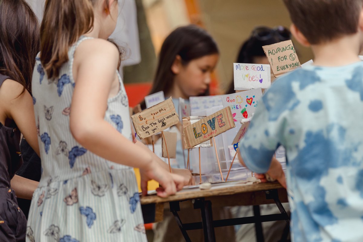Children gather around a table displaying colorful handmade signs with messages of love and positivity, such as "More good vibes," "Love always," and "Acupuncture for everyone."