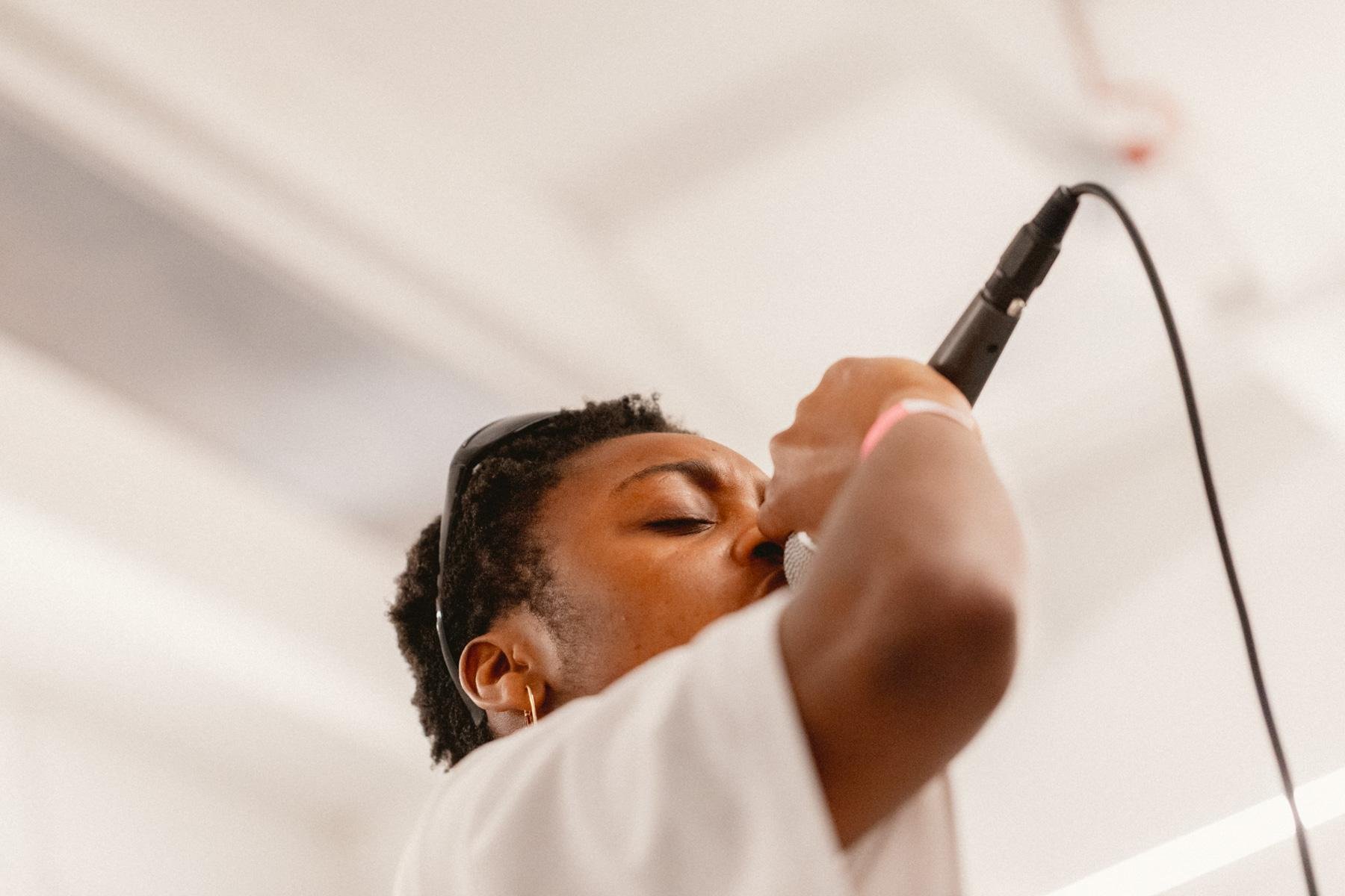 A woman singing into a microphone with her eyes closed, wearing headphones and a white top, standing in a room with white walls.