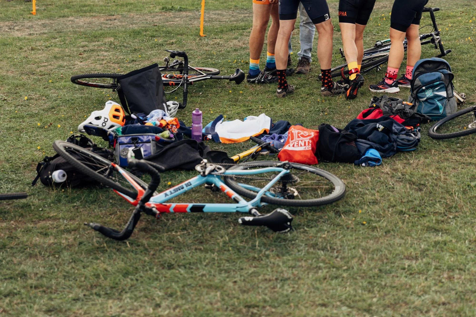 Various bicycles, helmets, water bottles, and sporting gear are scattered on the grass at an outdoor event. People are standing nearby, some wearing cycling clothing and socks.