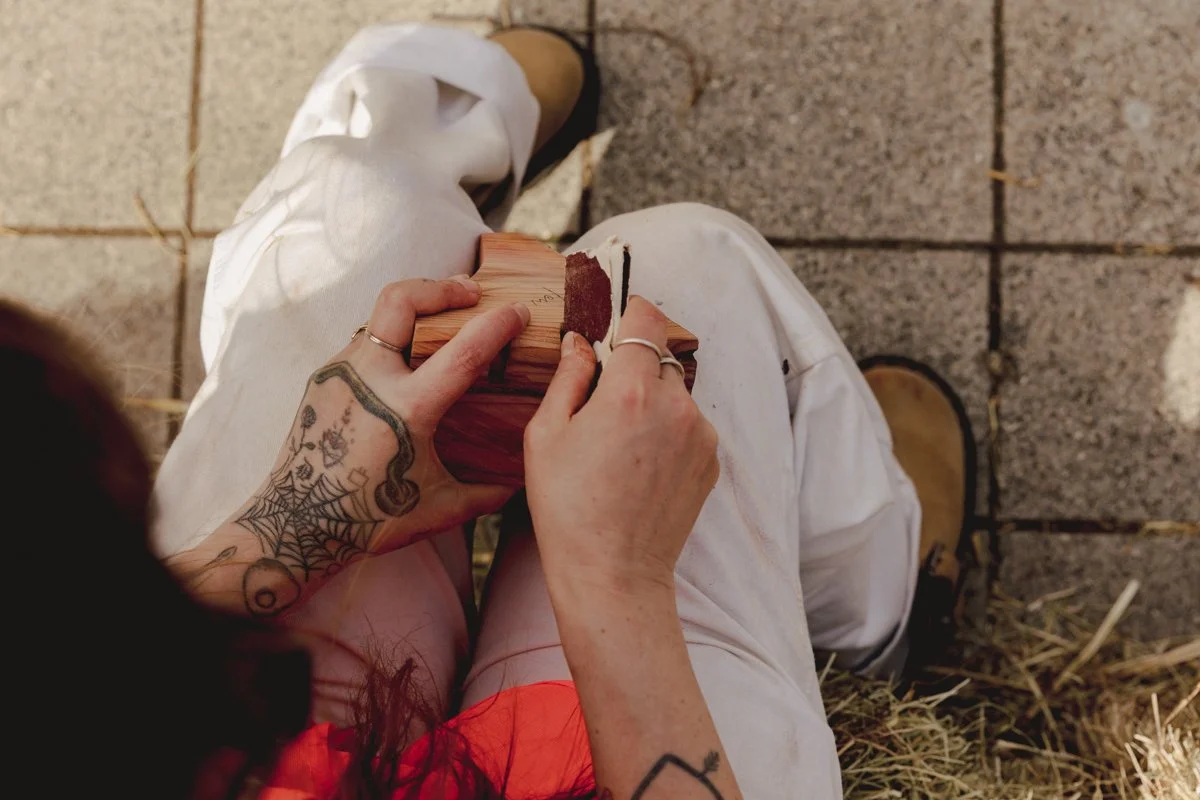 A person with tattoos on their hands and arms, sitting outdoors on a stone-tiled surface, holding a piece of wood and carving or shaping it with a small tool. They are wearing beige pants and tan shoes.