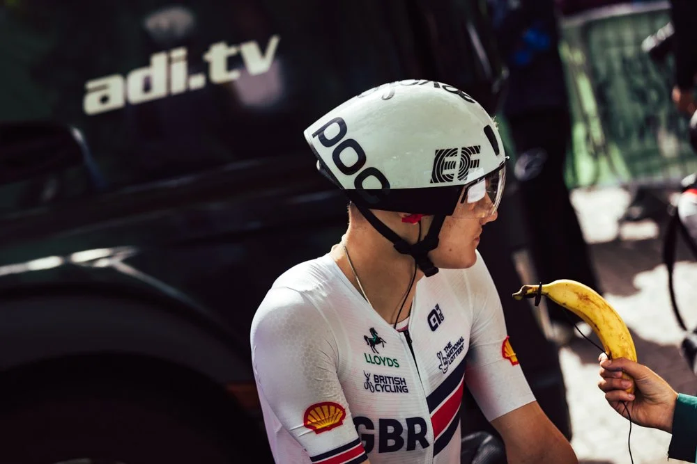 A female cyclist in a white GB racing suit and helmet is being offered a banana, with a person holding it on a stick in front of her. Vehicles and a bicycle are visible in the background.
