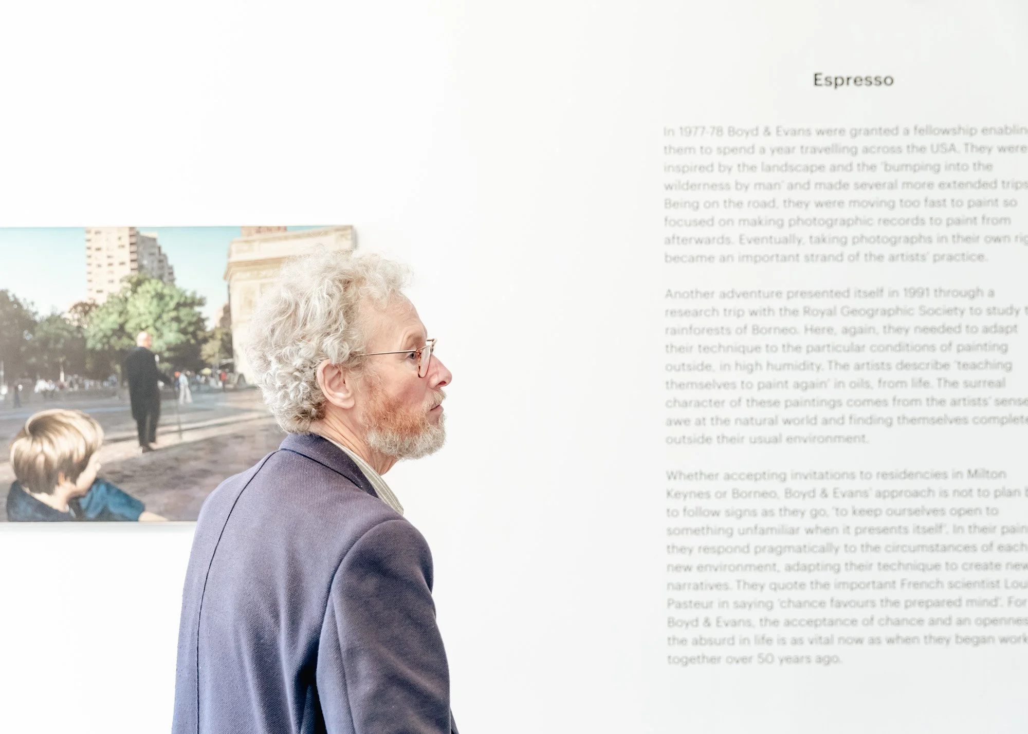 Older man with curly white hair and glasses, wearing a purple blazer, looking at an art display in a gallery.