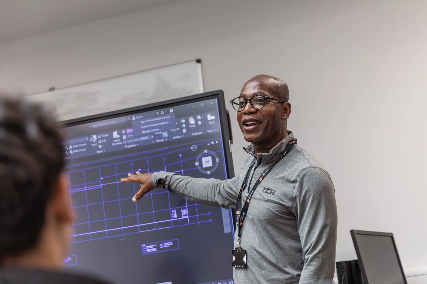 A man in glasses and a gray zip-up shirt is pointing at a large monitor displaying a CAD design or blueprint with a grid. He is smiling and standing in a classroom or conference room setting, with a whiteboard in the background.