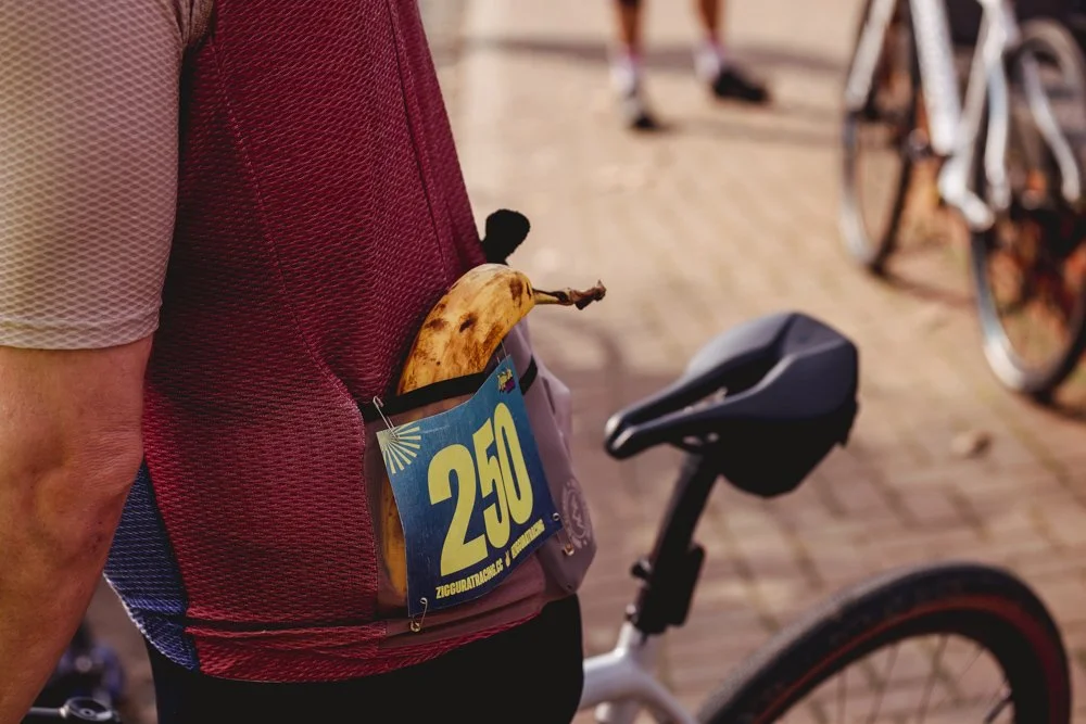 A close-up of a person wearing a pink and purple cycling vest, with a banana and a race bib number 250 tucked into the vest pocket, next to a bicycle.