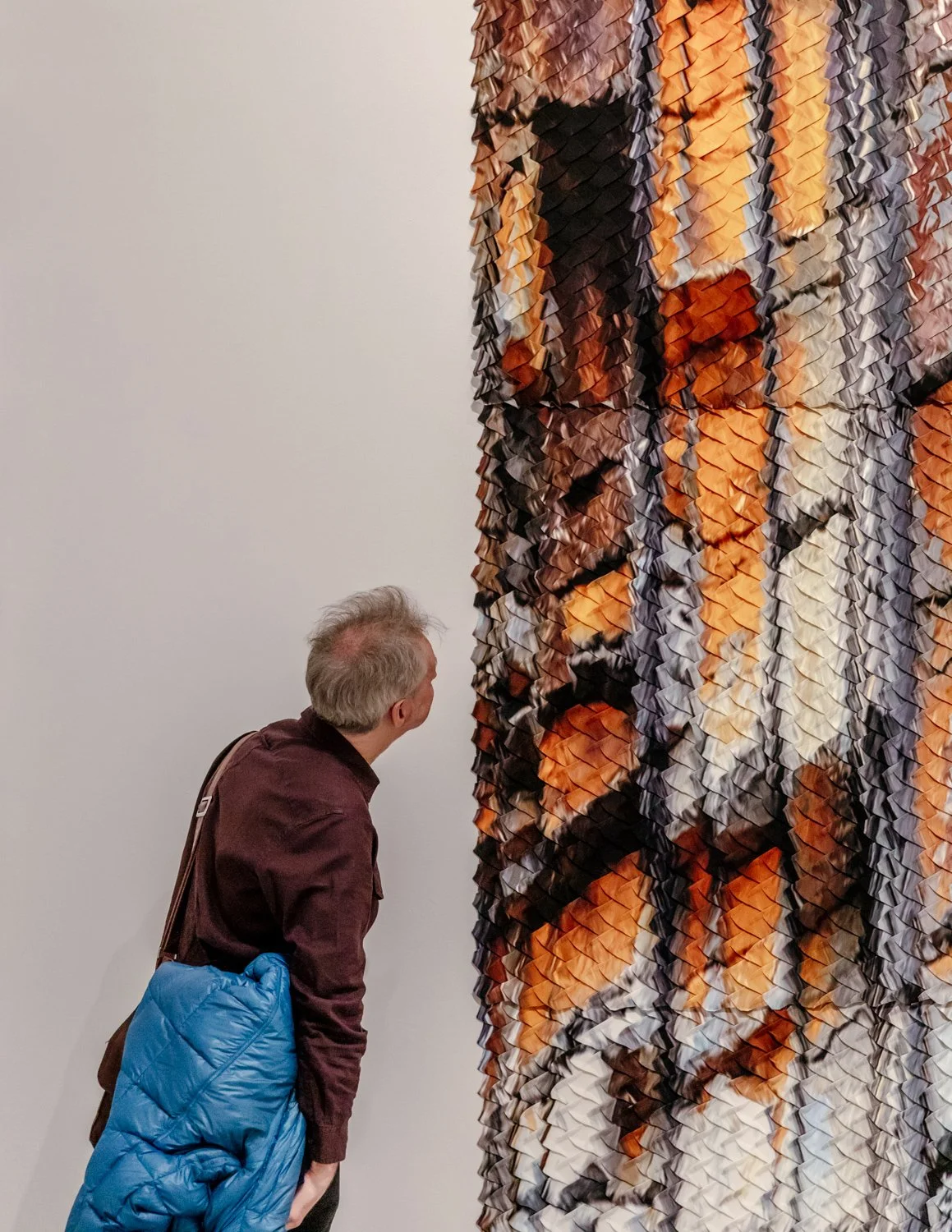 A person with gray hair and a maroon jacket is looking at a large, textured artwork hanging on a white gallery wall. The artwork is a large, sculptural piece composed of many layered, petal-like units in shades of orange, black, gray, and white, crea