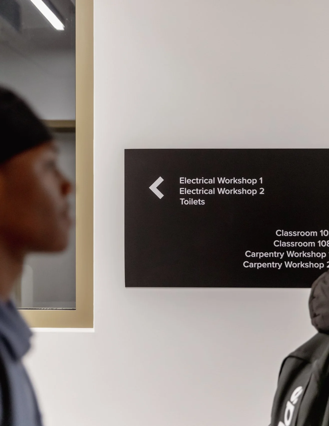People in a hallway near a wall sign pointing to electrical workshops, toilets, and classrooms.