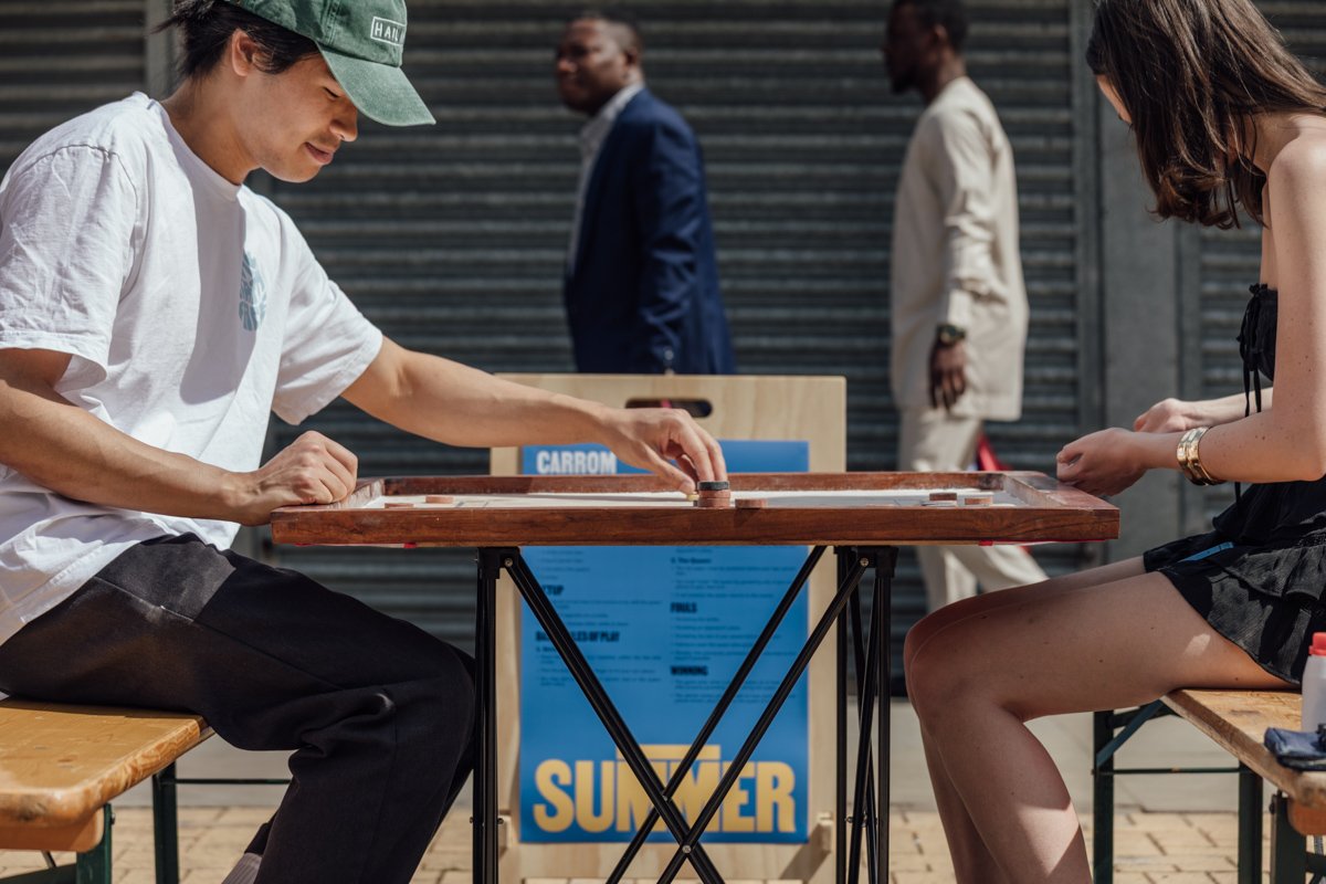 Two people seated at a wooden table playing shuffleboard outdoors, with two men walking in the background.