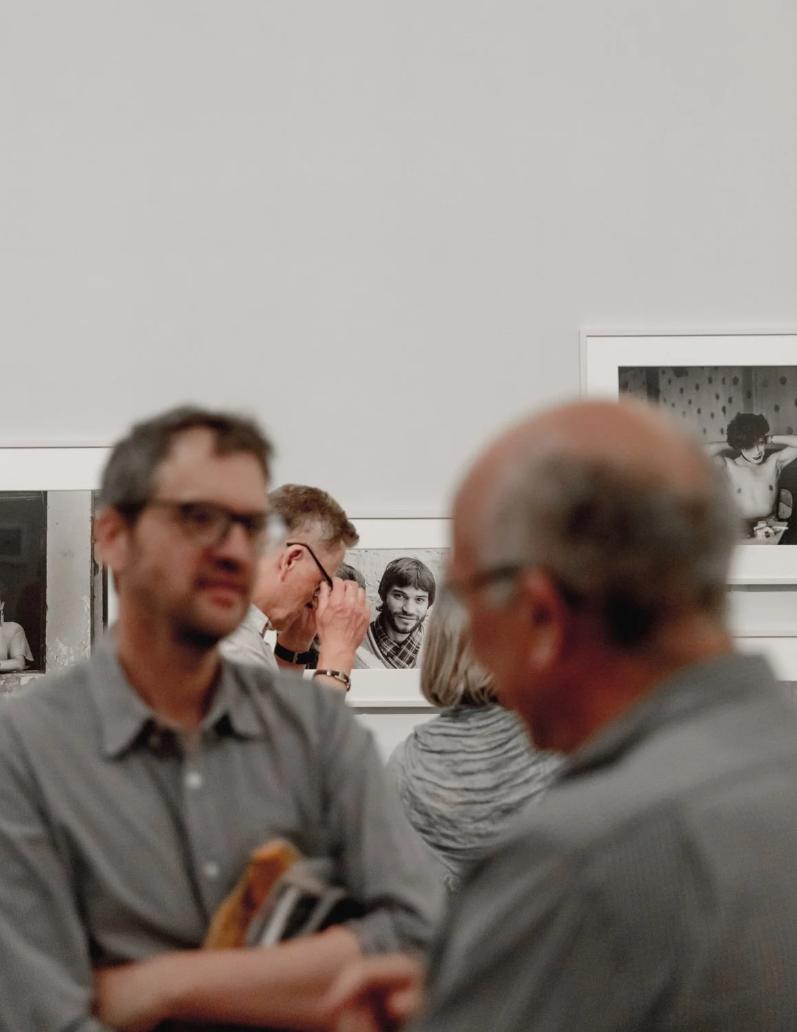 People at an art gallery, with photographs hanging on the white wall behind them, one person taking a photo, and others engaged in conversation.