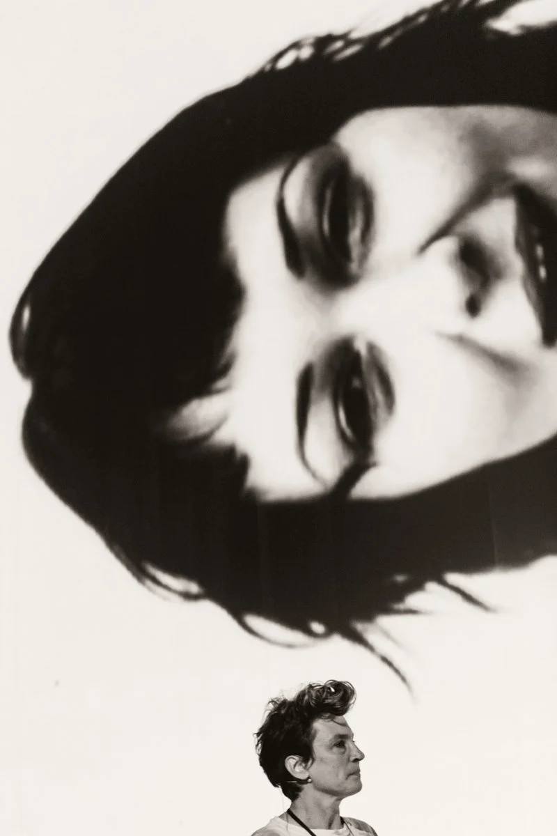 A woman with short, wavy hair standing in front of a large screen displaying a close-up of her own face.