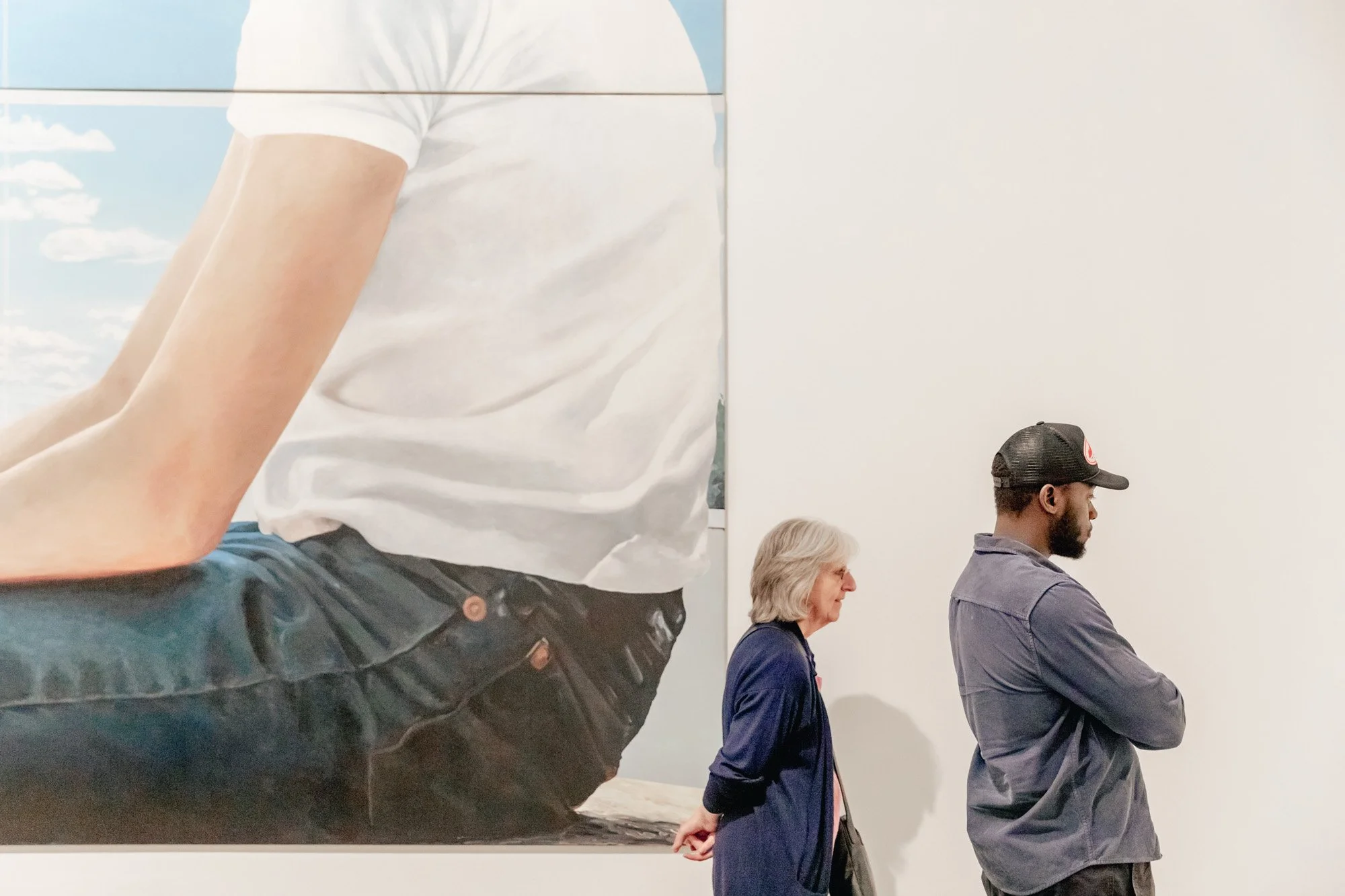Two people standing in front of a large painting in an art gallery. The painting depicts a person wearing a white shirt and black pants, with a bright blue sky background.