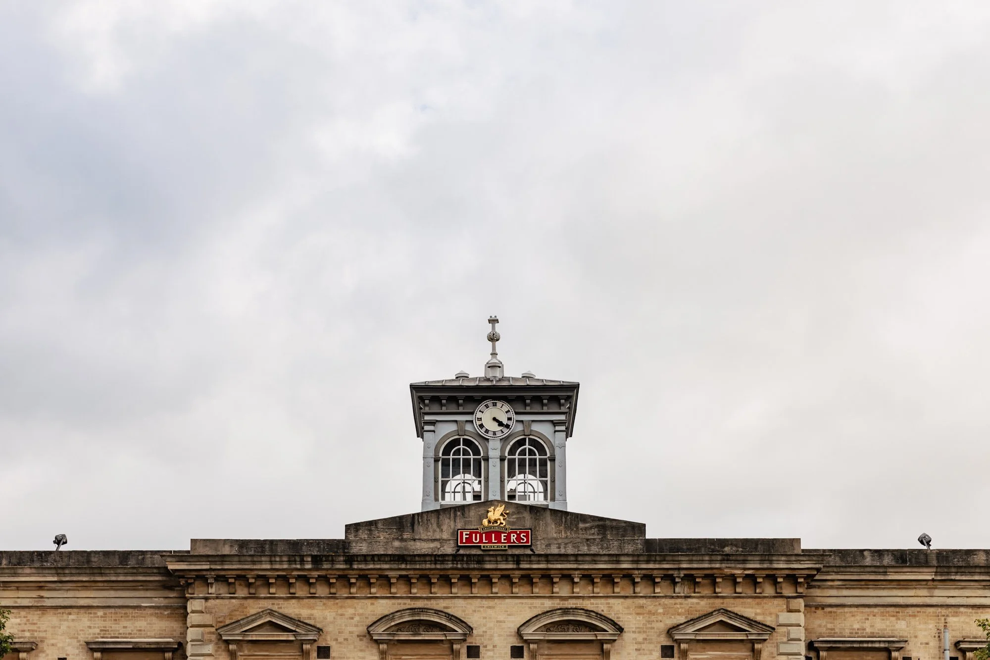 A historic building with a clock tower featuring two large arched windows and a clock showing 11:40. The building has a sign that reads 'Fuller's' with a lion logo above it. The sky is cloudy with a few spotlights on the roof.