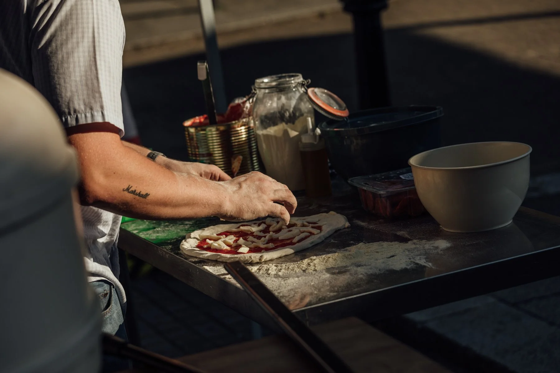 Person preparing a pizza on a floured surface outdoors, surrounded by bowls and containers of ingredients.