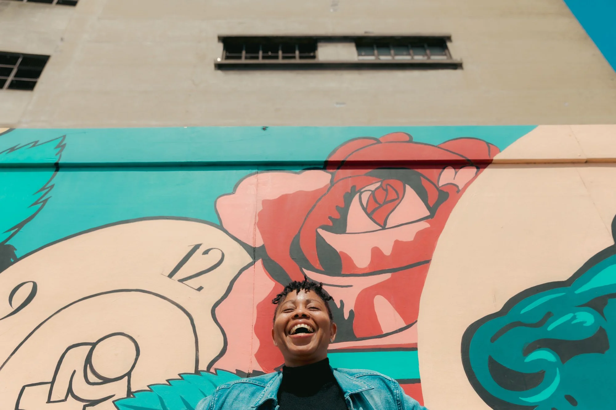 A woman with short curly hair smiling widely, standing in front of a colorful mural with a large rose and a clock.