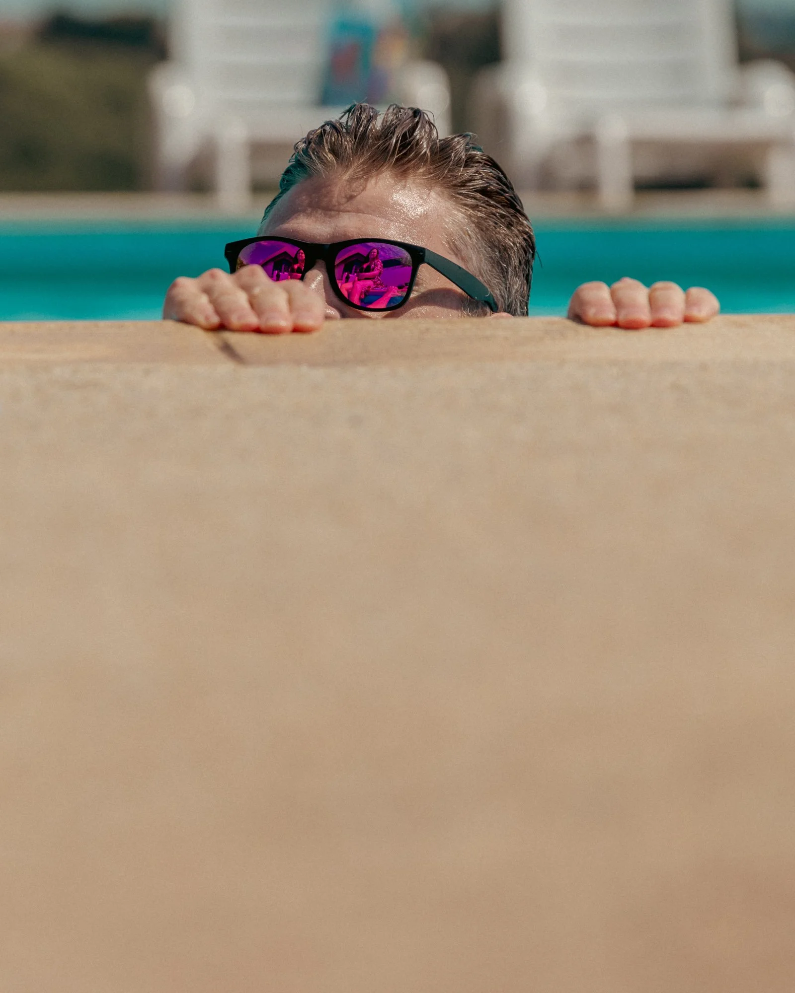 Person with sunglasses peeking over the edge of a poolside deck, with pool water and lounge chairs in the background.