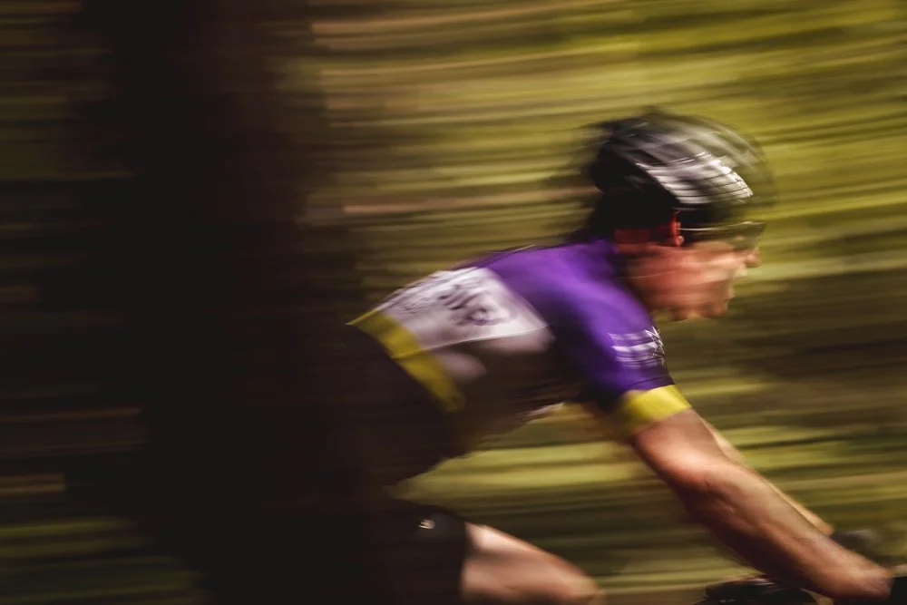 A cyclist wearing a purple, black, and yellow jersey, black helmet, and sunglasses riding fast through a wooded area with blurred background.