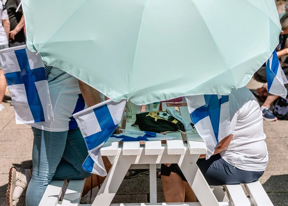 People sitting at a white picnic table under a large light green umbrella, with historic Greek flags attached to the table. The scene appears to be outdoors on a sunny day.