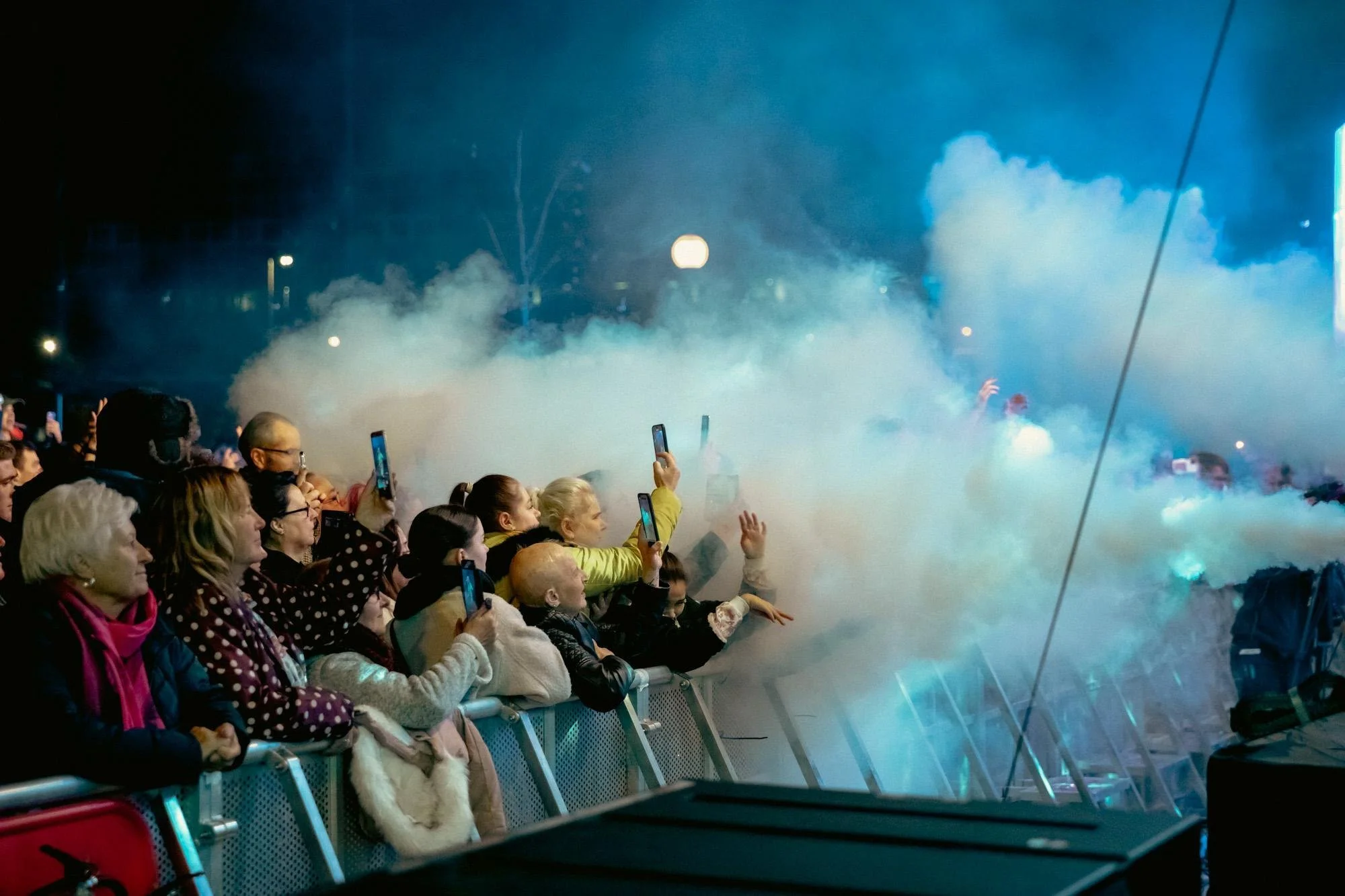 Crowd of people in winter clothing taking pictures and reaching toward a stage while smoke or fog effects create a misty atmosphere at night during an outdoor event.