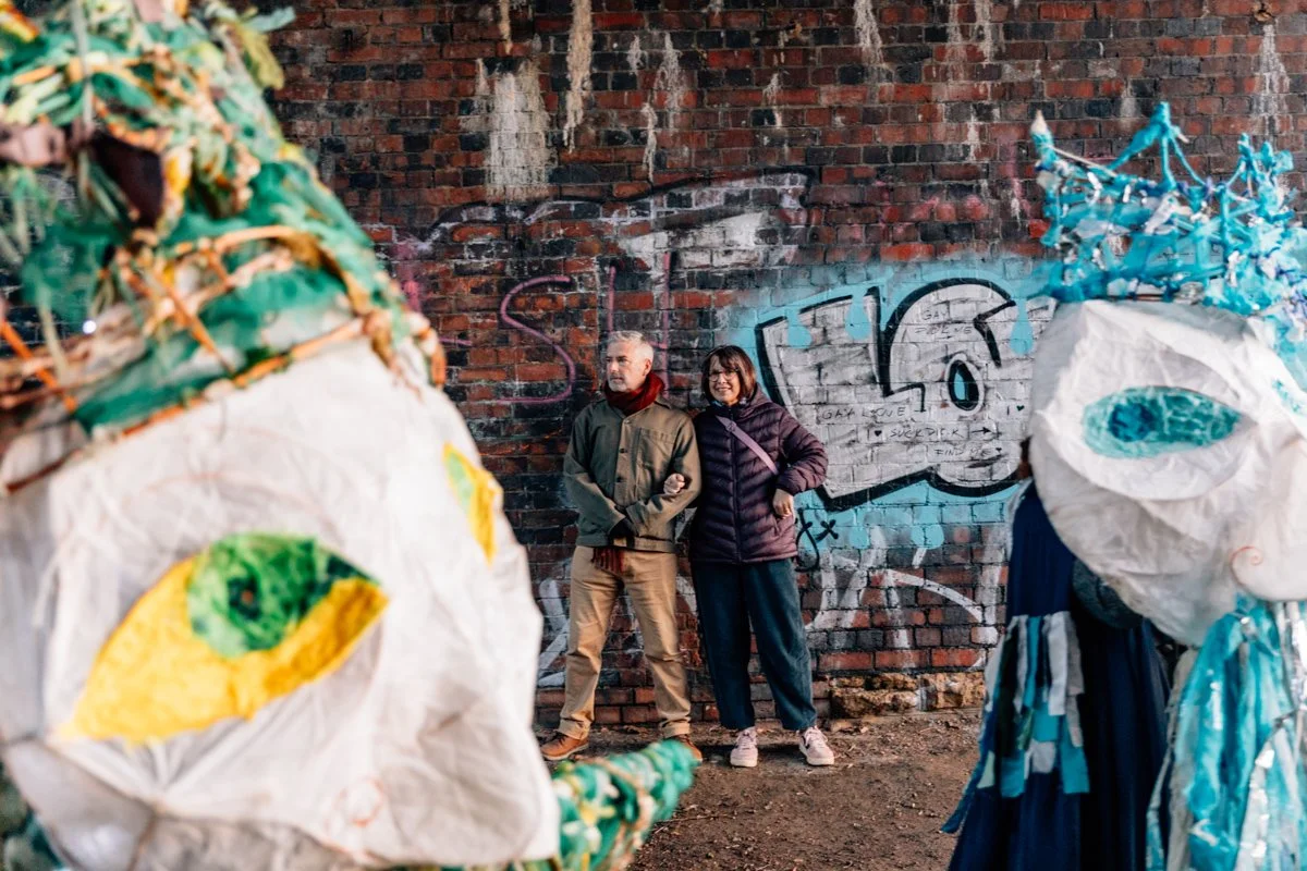Two people standing against graffiti-covered brick wall, with large colorful lantern sculptures in the foreground.