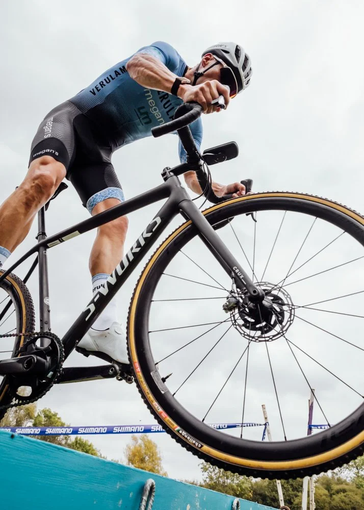 A cyclist wearing a helmet and sports gear, riding a black mountain bike on a trail with a blue barrier in front and trees in the background.