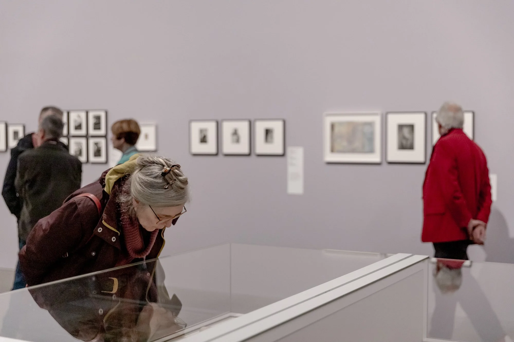 Museum visitors observing framed artwork on a gallery wall, with one person looking into a glass display case.