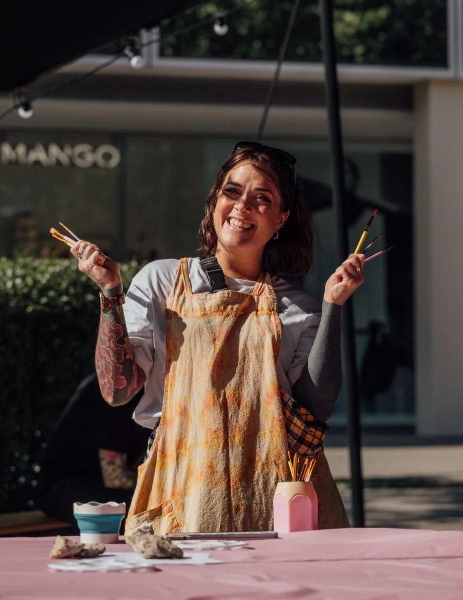 Young woman smiling outdoors, holding paintbrushes, wearing a multicolored apron and tattoos on her arms, standing behind a table with art supplies.