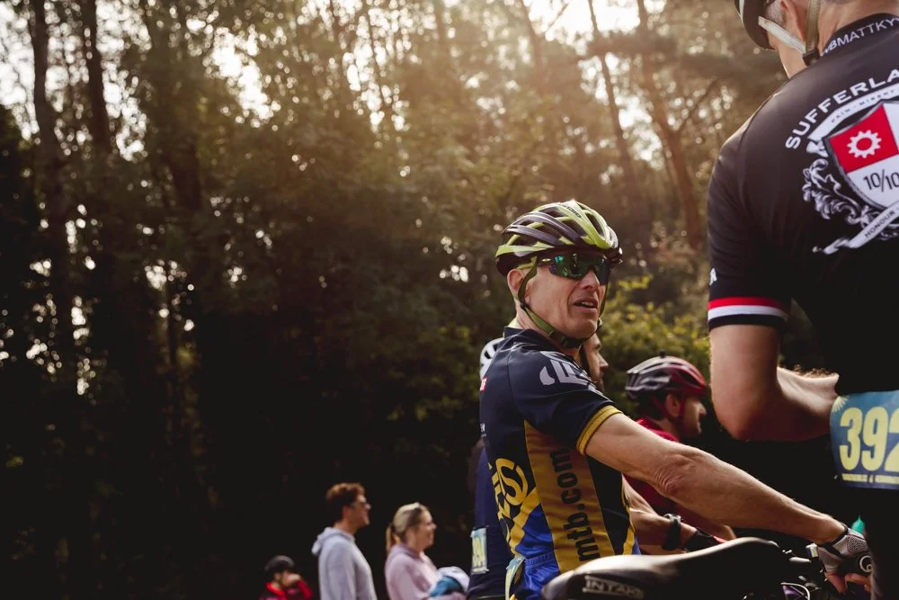 Cyclists, including an older man wearing sunglasses and a helmet, preparing for a ride outdoors with trees and sunlight in the background.