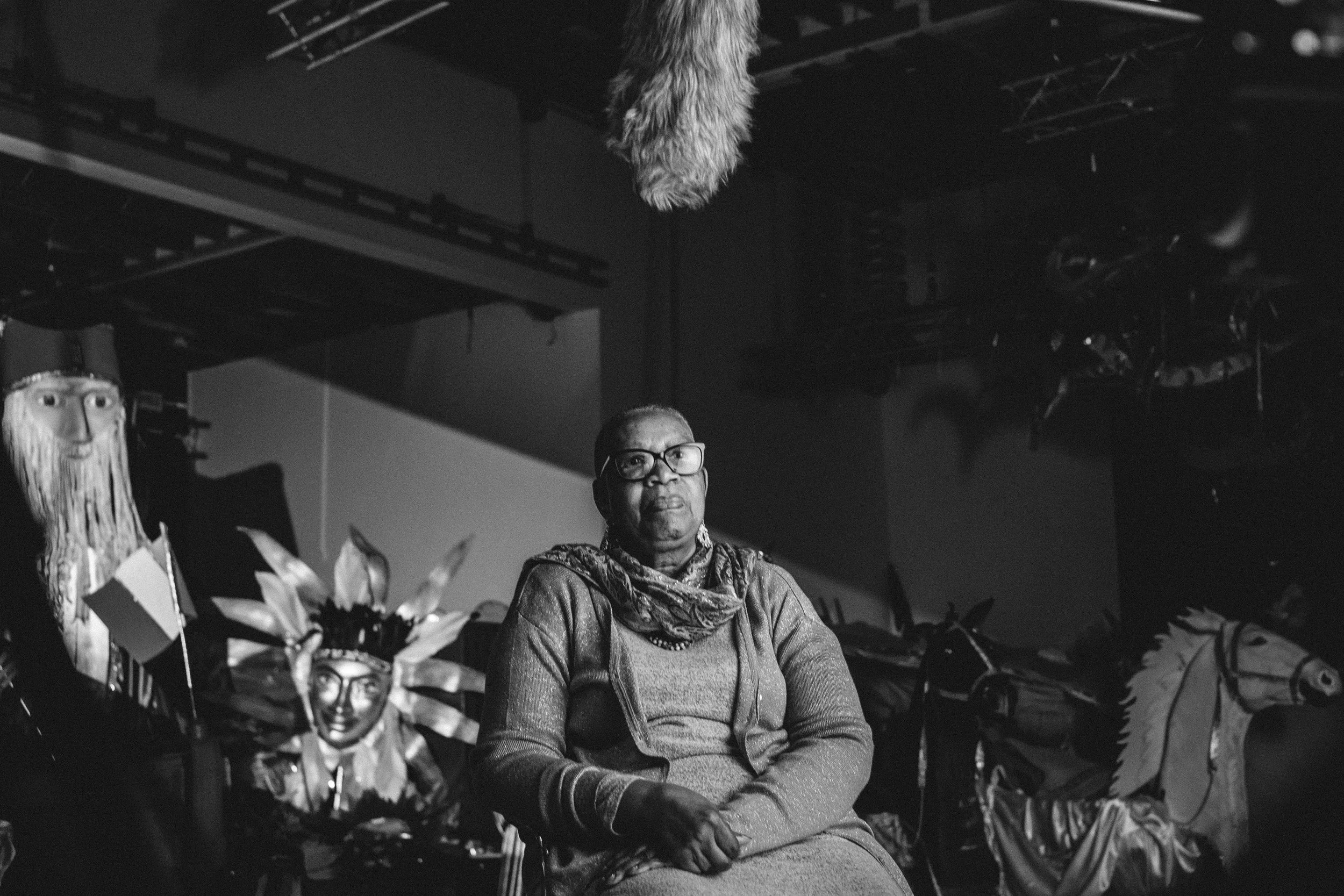 A woman with glasses sitting among various colorful decorations, including a clown face with feathers, a large wooden horse, and other cultural artifacts, in a dark setting.