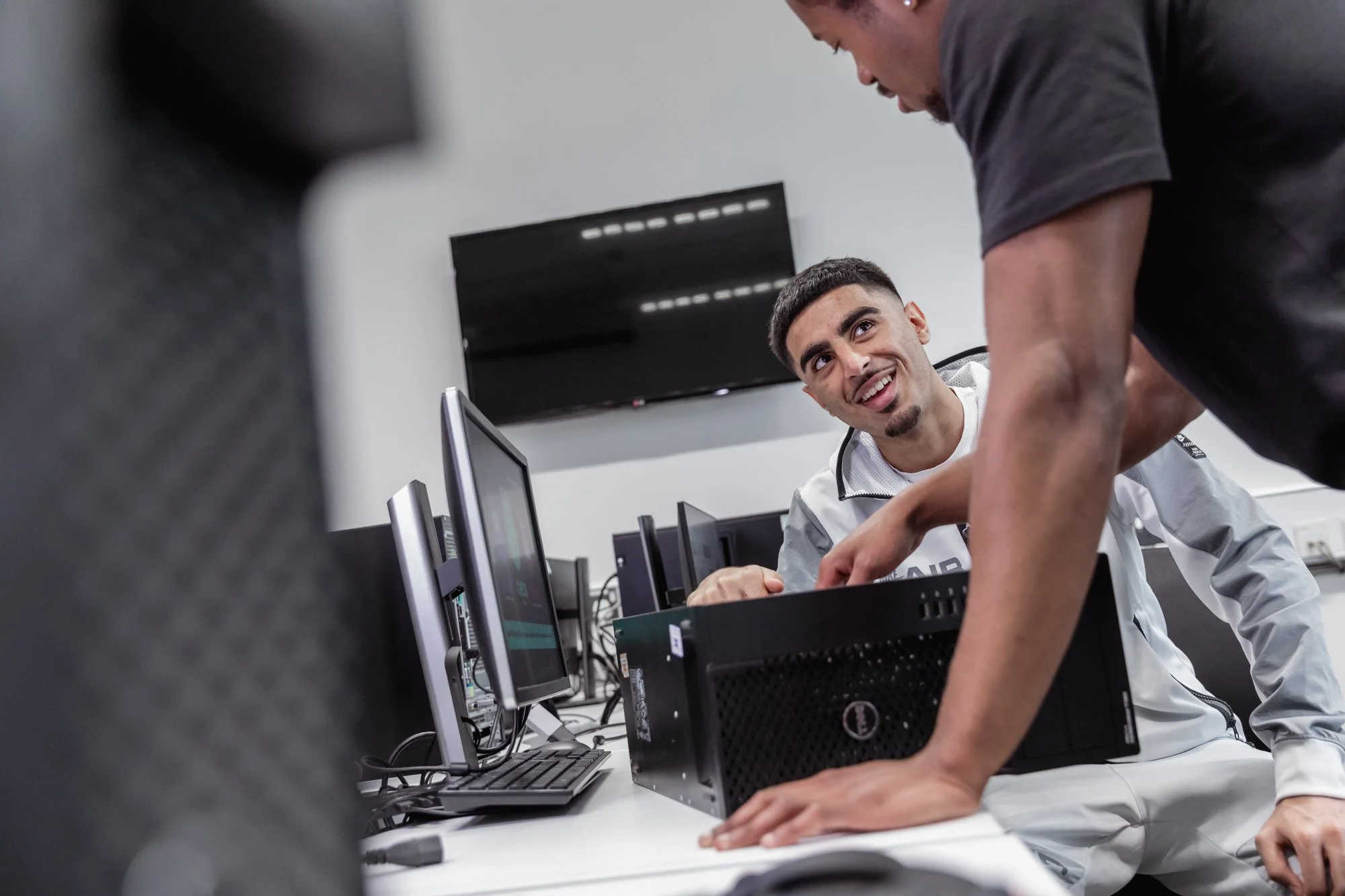 Two men working on a computer setup in an office, one sitting at the desk and the other leaning over, engaging in conversation.