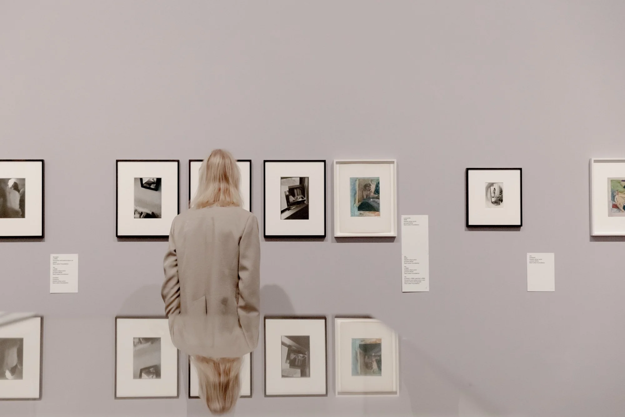 A person with blonde hair wearing a beige jacket looking at framed artwork on a gray gallery wall. The artwork includes photographs and paintings. There are informational plaques next to some frames.