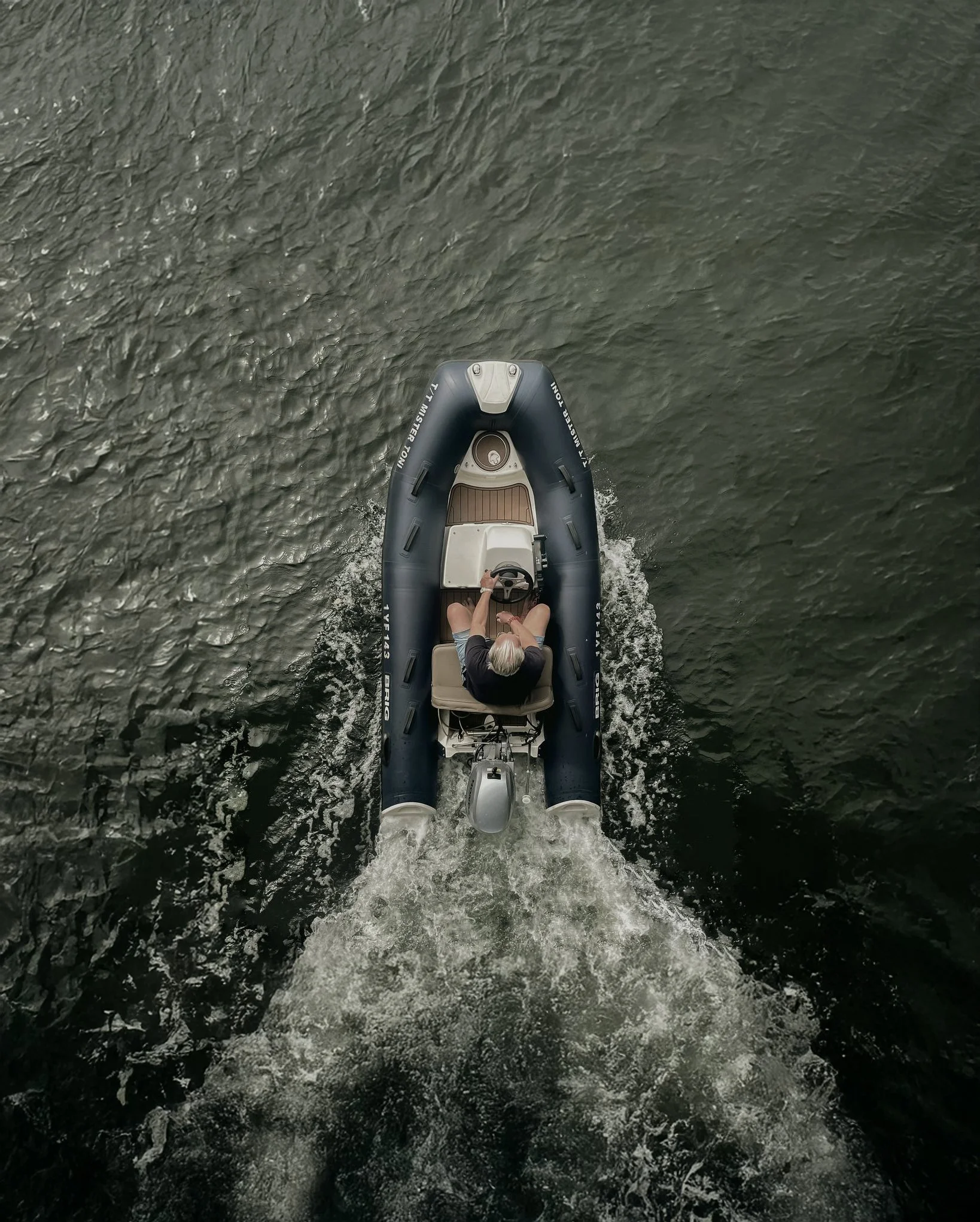 A person with gray hair driving a small inflatable boat with an outboard motor on a body of water, seen from above.