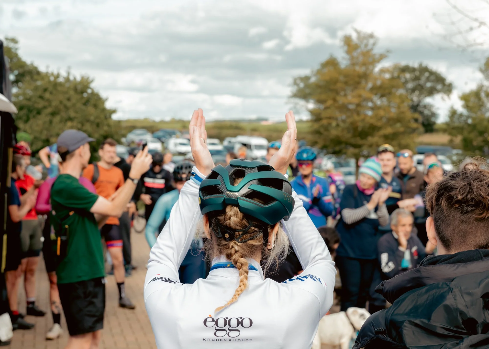 A group of people gathered outdoors, many wearing cycling gear and helmets, with one woman in the foreground holding her head with hands raised, participating in a team event or race.
