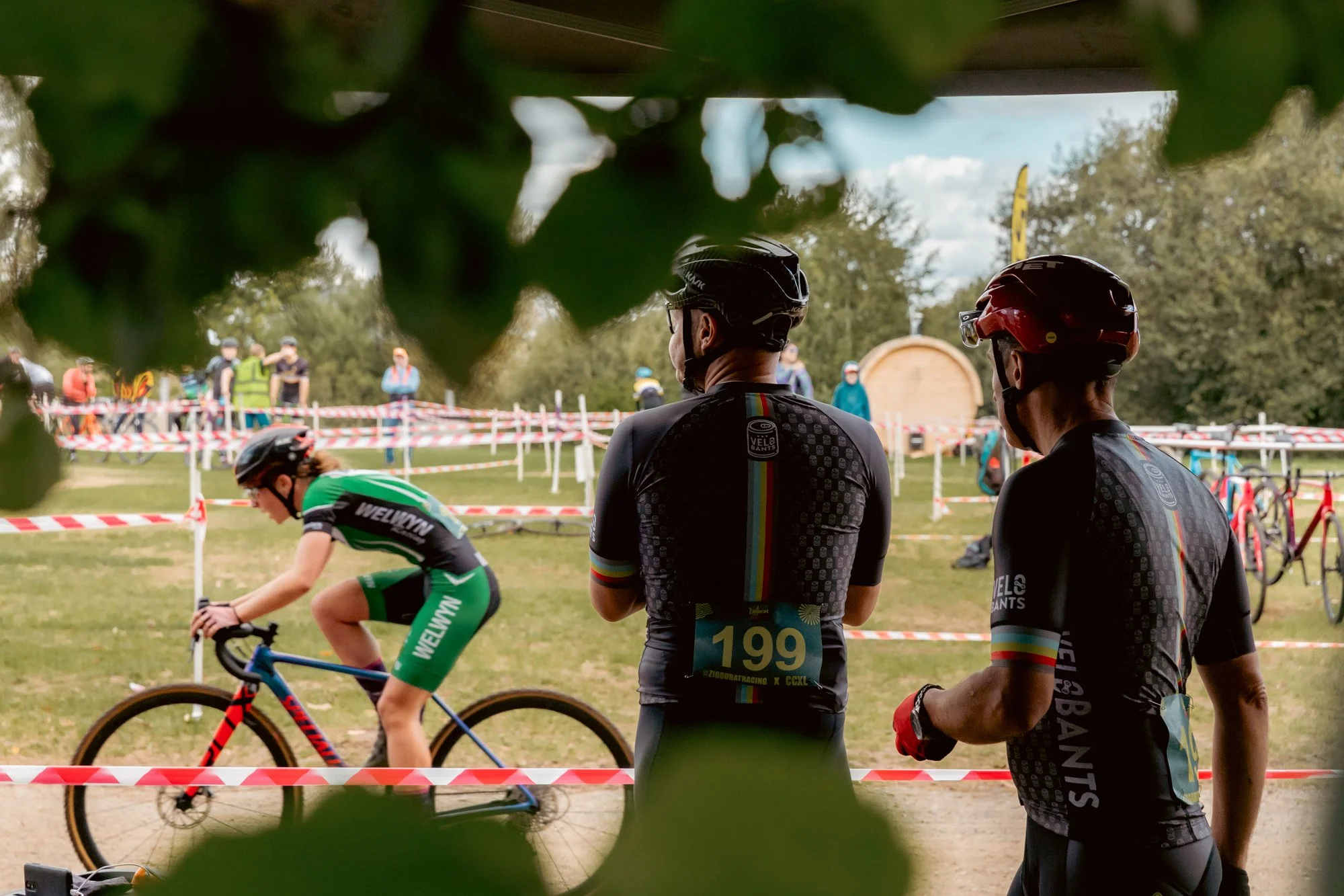 Cyclist racing past two officials or coaches watching from the sidelines during a mountain biking competition, with a dirt trail, red and white barrier tape, and spectators in the background.