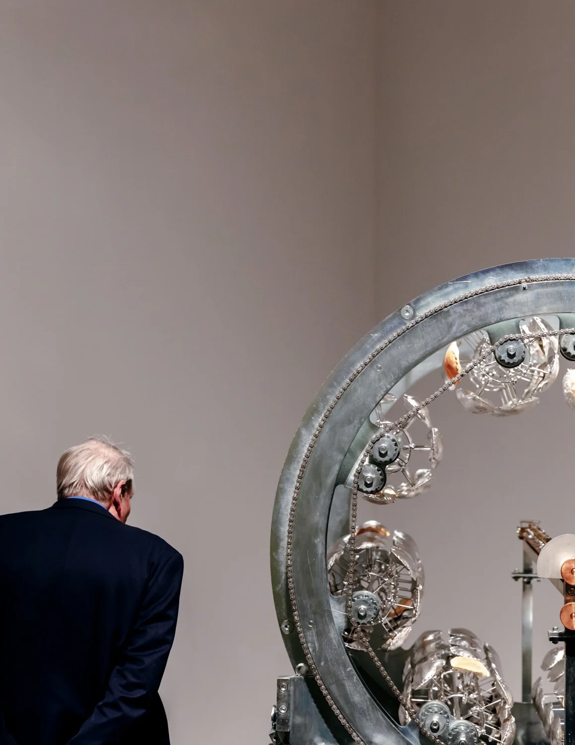 An older man with white hair wearing a dark blazer is gazing at a large metallic scientific instrument with multiple glass and metal components.