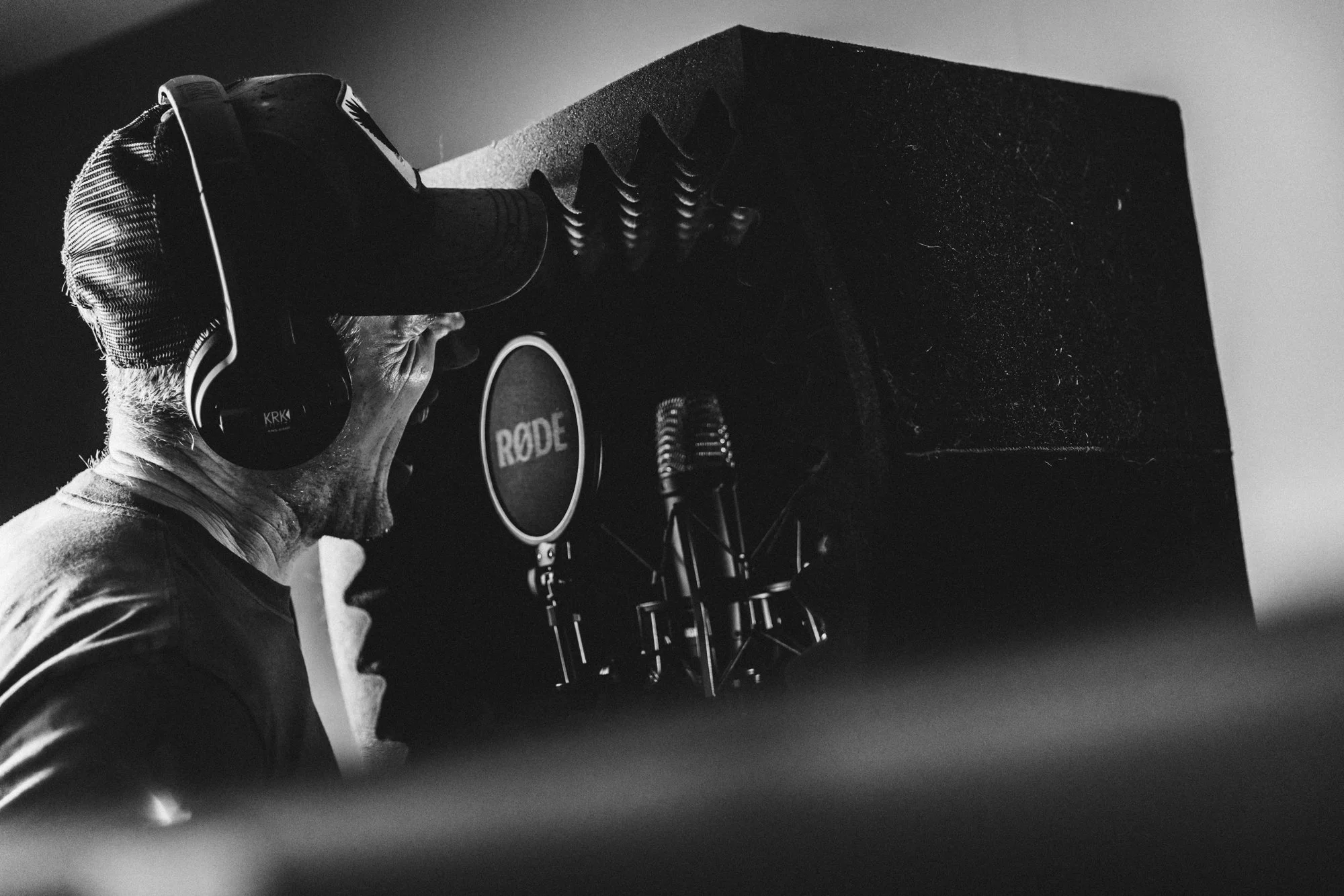 A man singing into a microphone in a recording studio, wearing headphones, with a large speaker behind him.