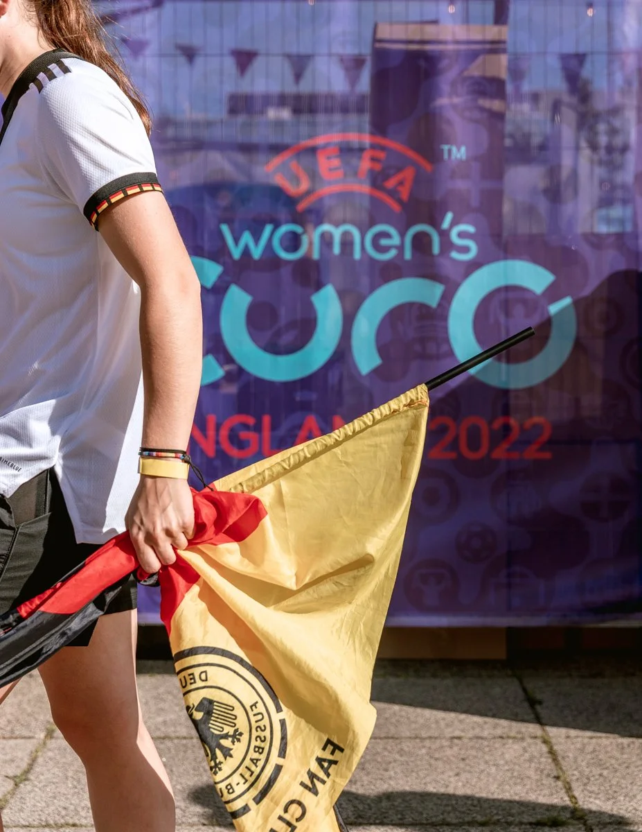 A person holding a yellow and red flag in front of a blue banner that reads "UEFA Women's Euro England 2022."