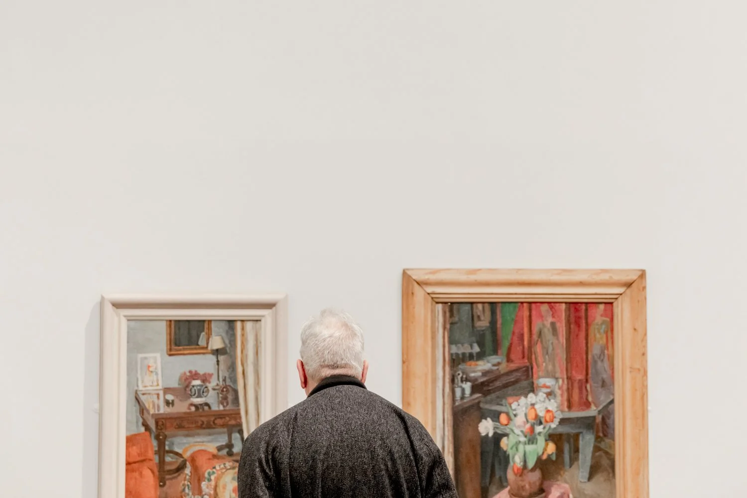 An elderly man with gray hair wearing a dark coat is standing in an art gallery, looking at two framed paintings on a white wall.
