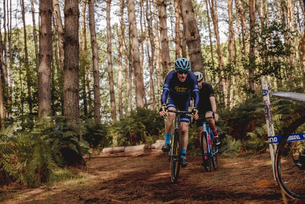 Two mountain bikers riding on a dirt trail through a forest with tall trees and green ferns.