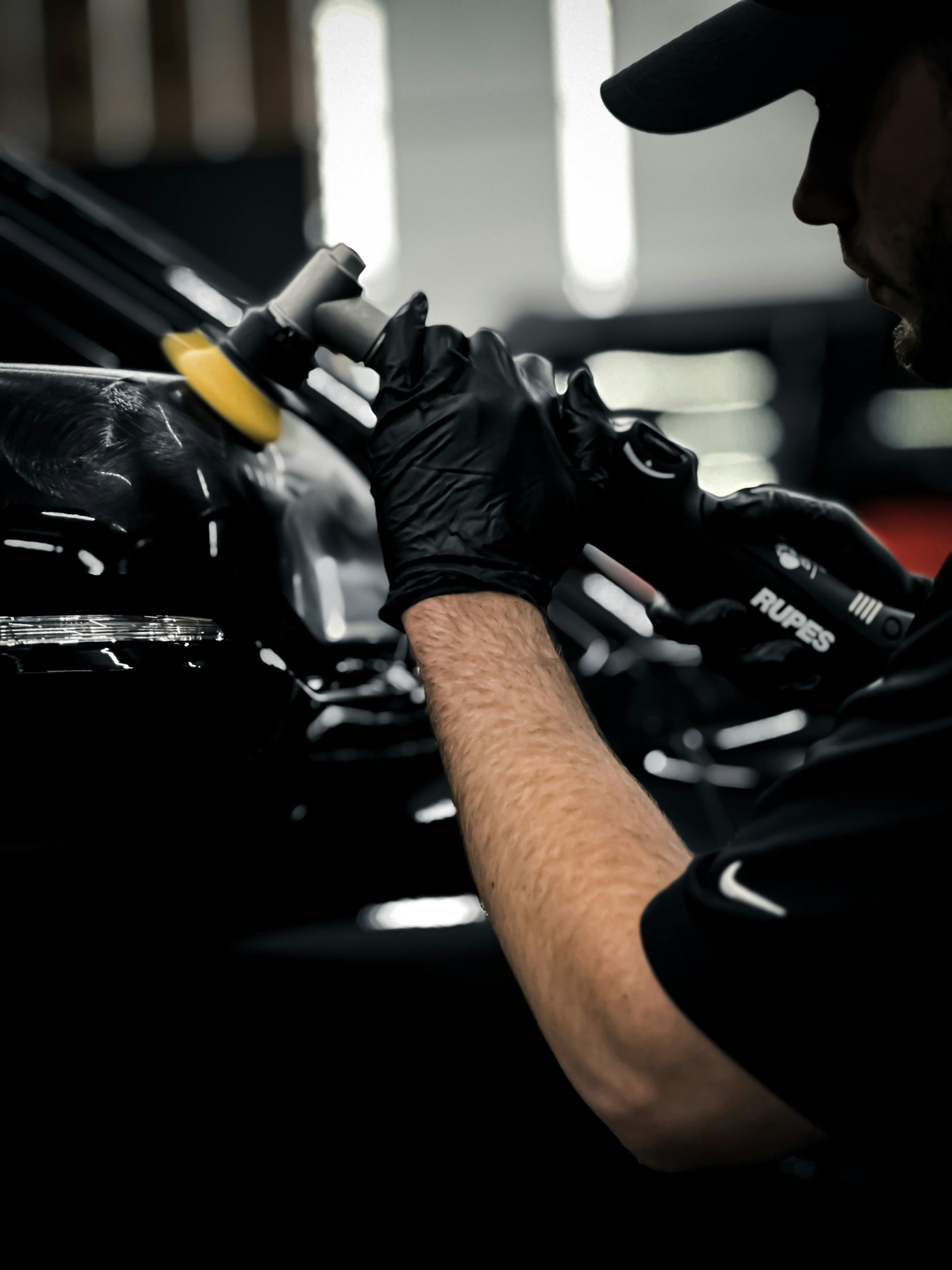 A person wearing a black cap and black gloves polishing a black car with a yellow polishing pad in a garage.