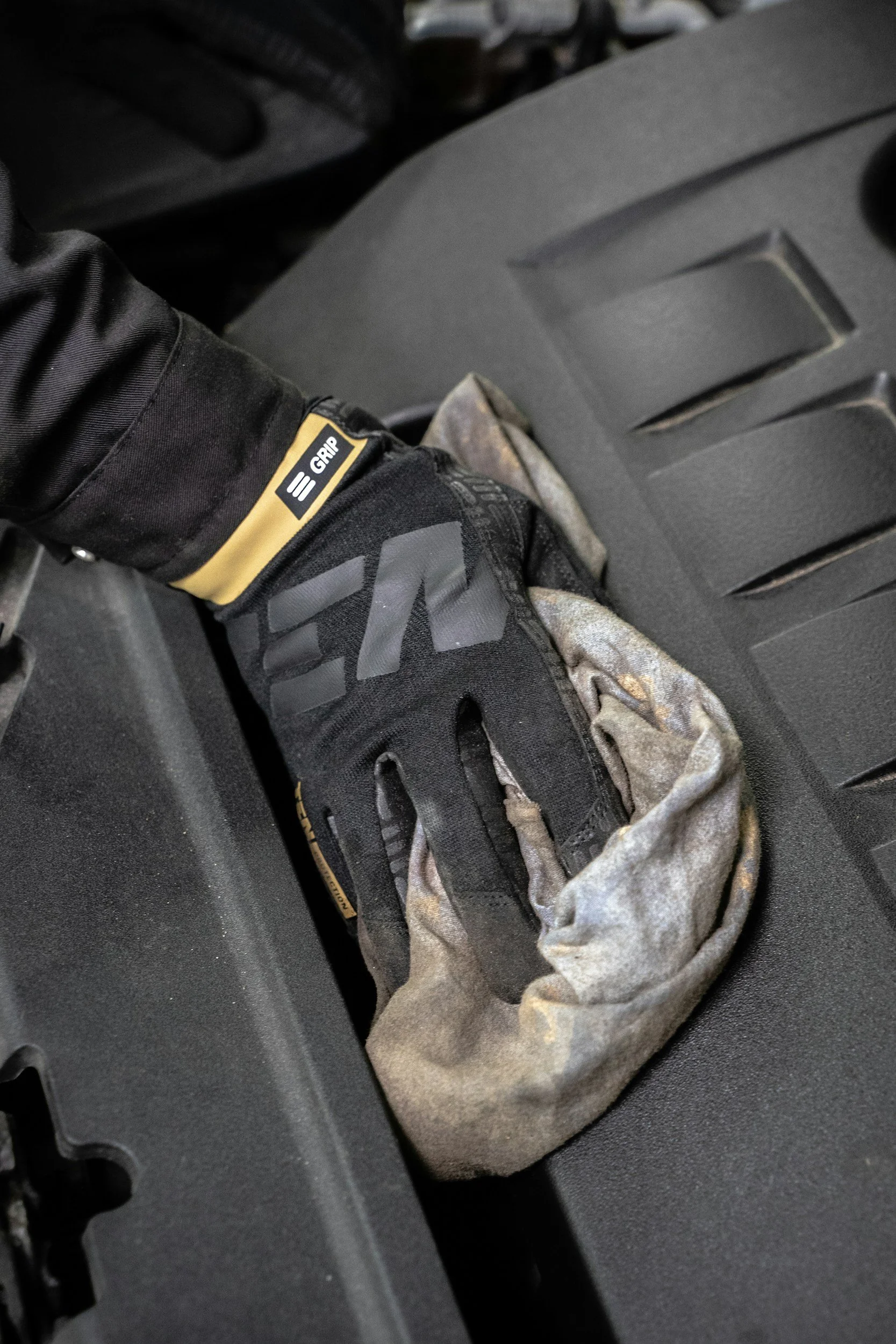 A gloved hand resting on a black surface, with part of a vehicle engine visible.