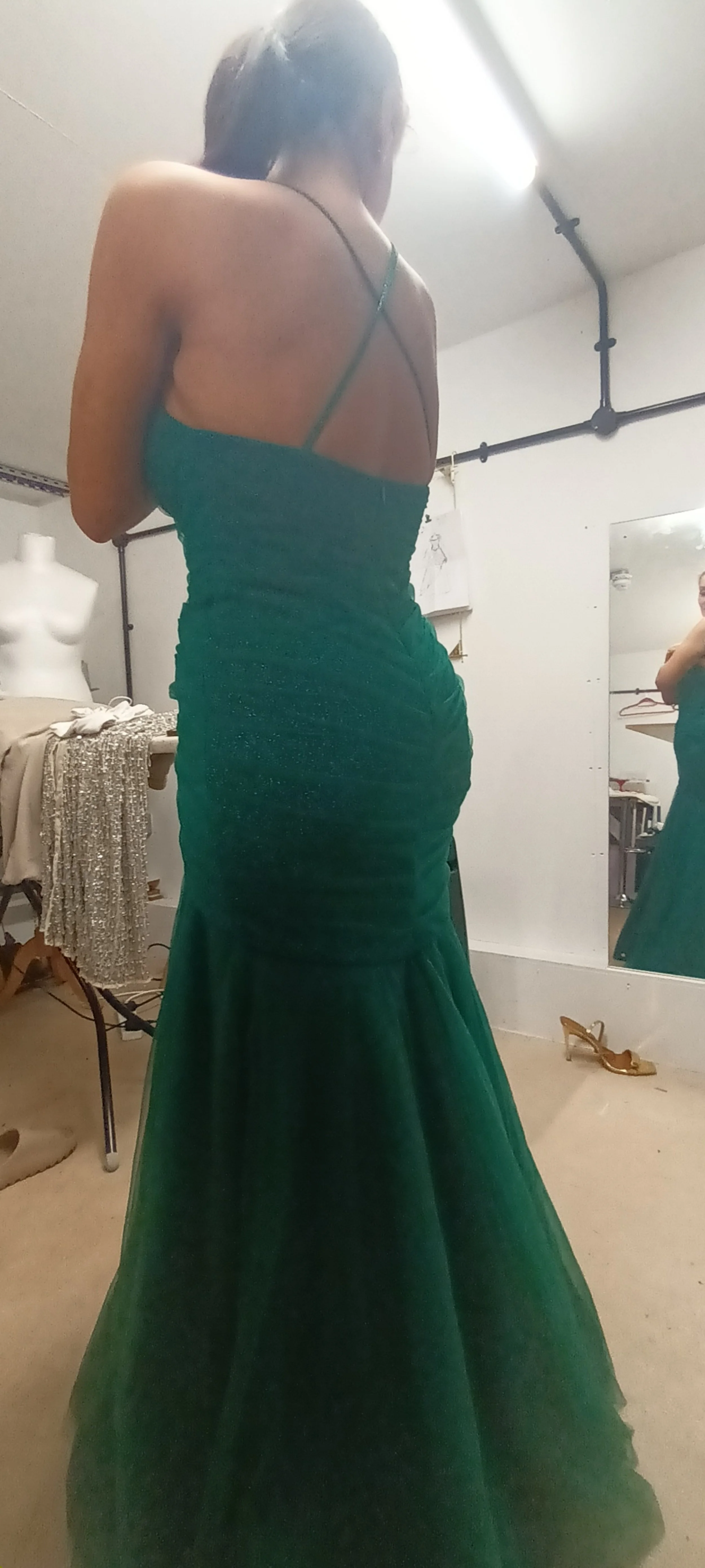 A woman in a green, strapless gown with ruffling details, standing in a fitting room, taking a mirror selfie.