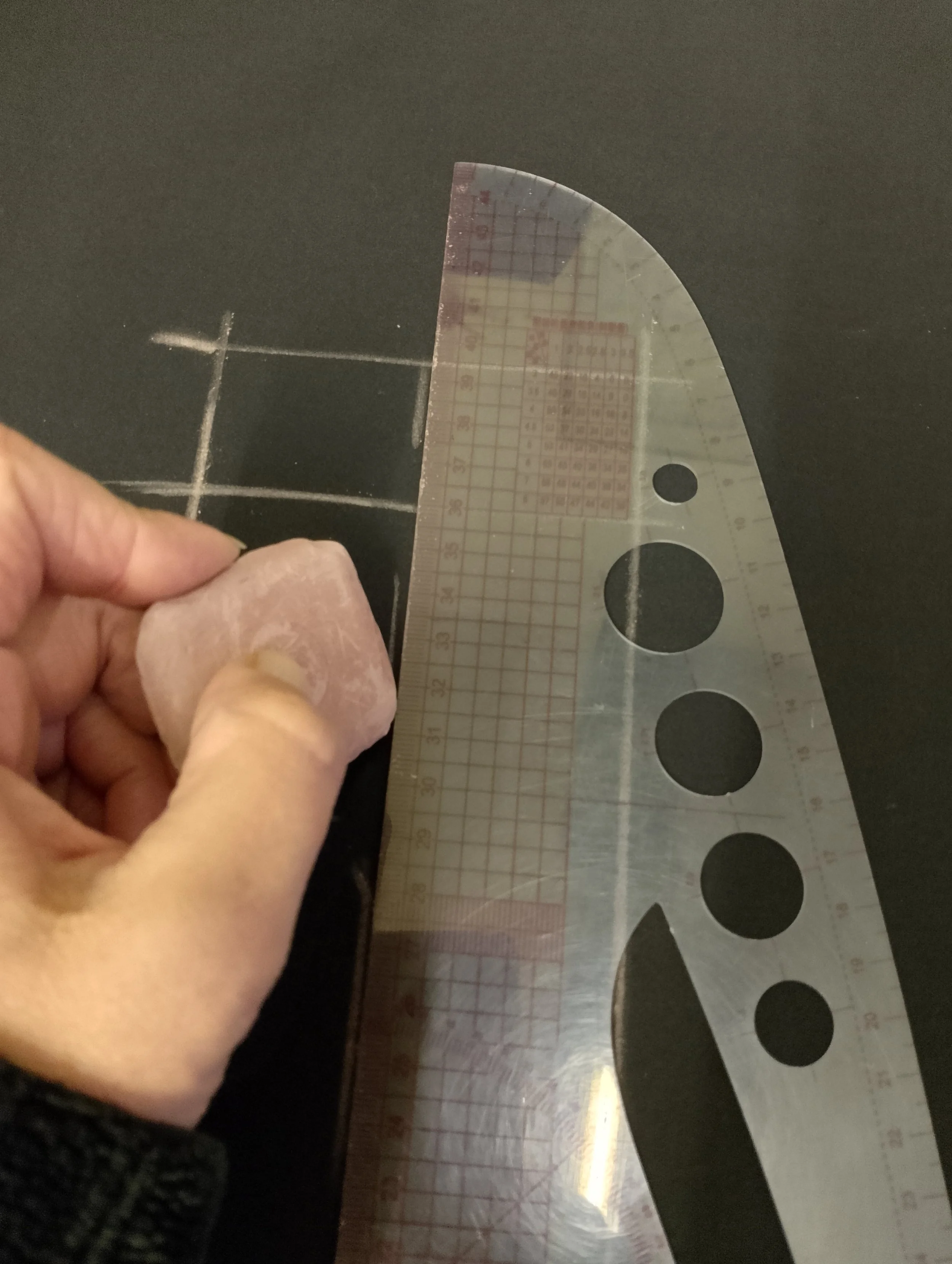 Person holding a piece of pink rock or mineral with their left hand, near a transparent geometric protractor on a black surface with white grid lines.