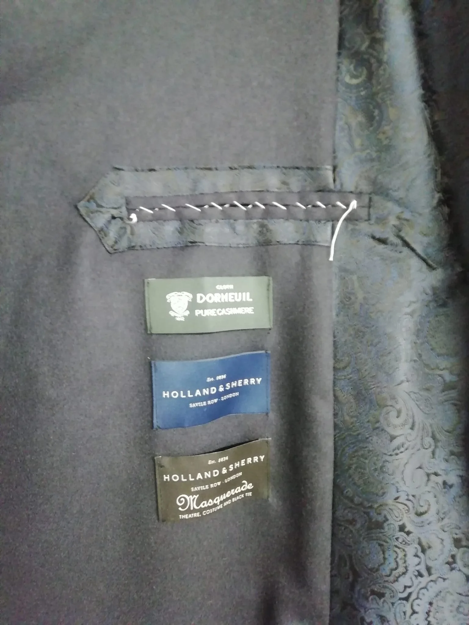 Internal view of a bespoke men's jacket in progress, featuring a hand-finished jetted pocket secured with basted cross-stitching and authentic labels for Dormeuil Pure Cashmere and Holland & Sherry fabrics, hand-crafted in the Elena Milani studio.
