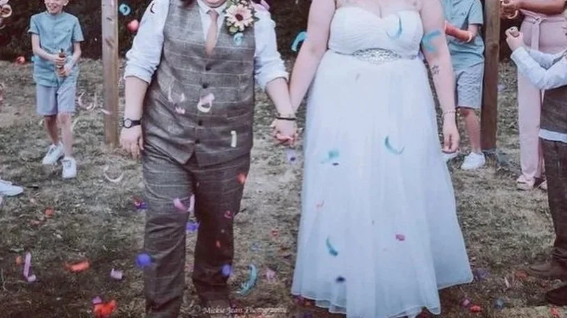 A same-sex couple holding hands at a wedding ceremony outdoors, with guests celebrating and confetti falling.
