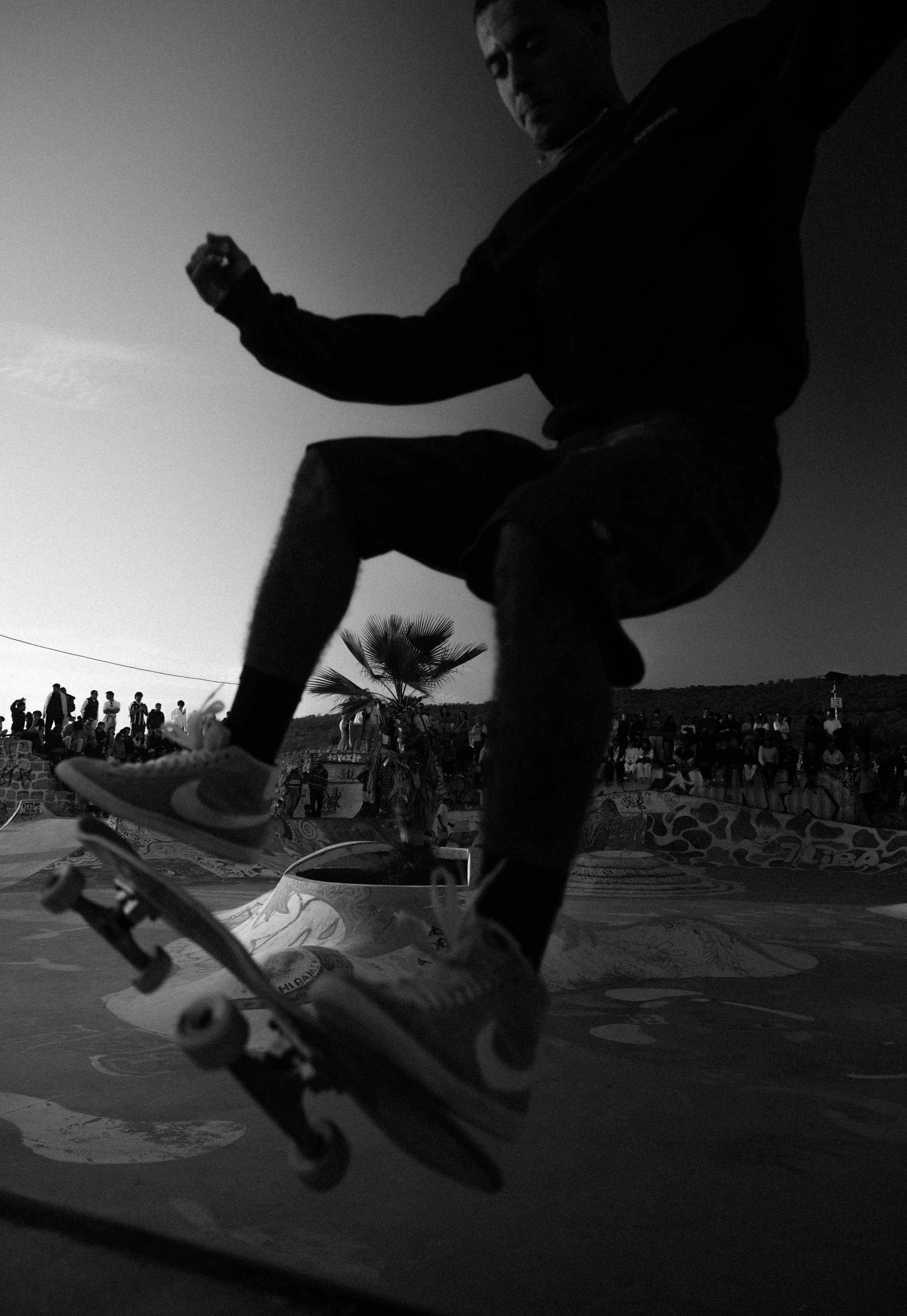 Skateboarder in mid-air performing trick at skatepark during sunset, crowd watching in background, black and white photo.